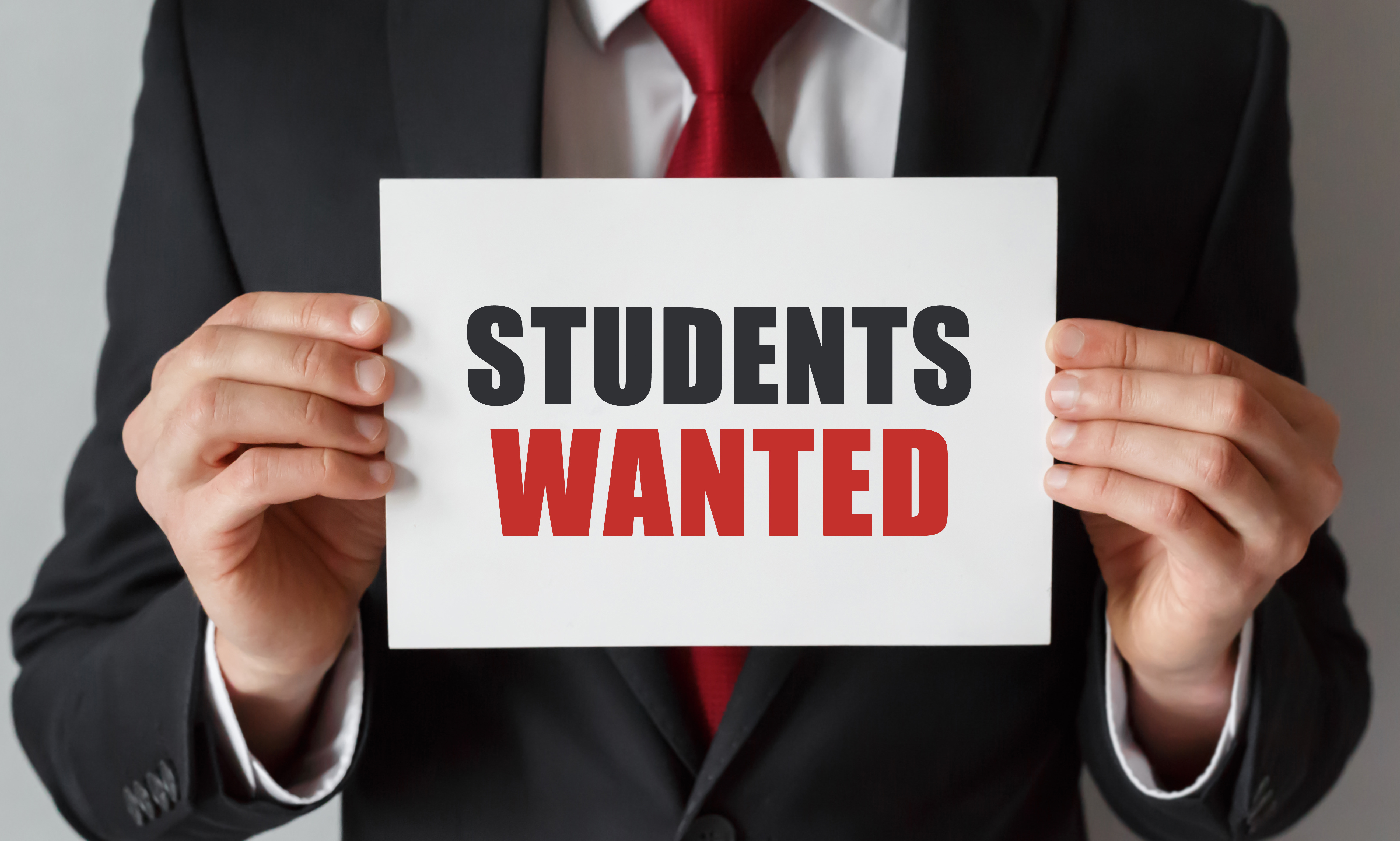 Person in a suit holding a sign that says, Students Wanted