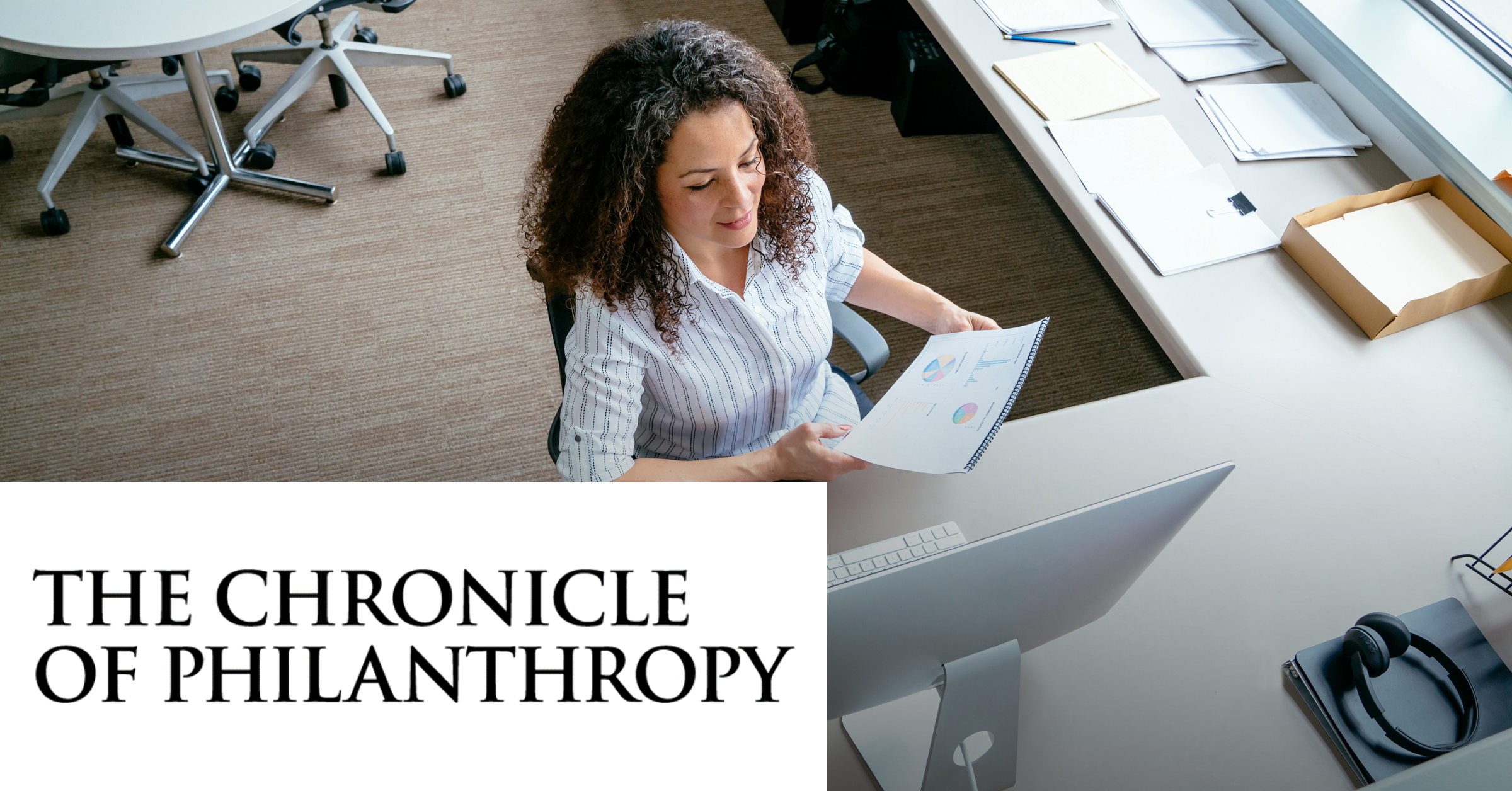 Image of a woman working at a office in front of the computer while reading a physical document. On the lower left corner is placed the Chronicle Of Philanthropy black logo on a white horizontal rectangle.