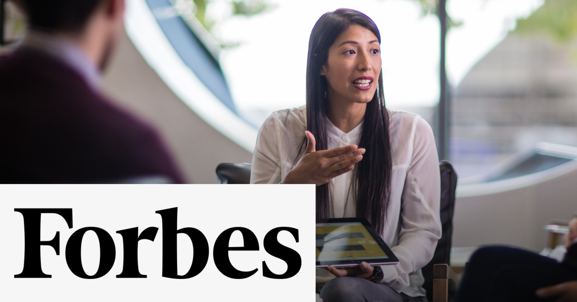 Image of a woman during a speech using an electronic tablet to show her presentation. On the lower left corner the Forbes black logo is placed on a white rectangle.